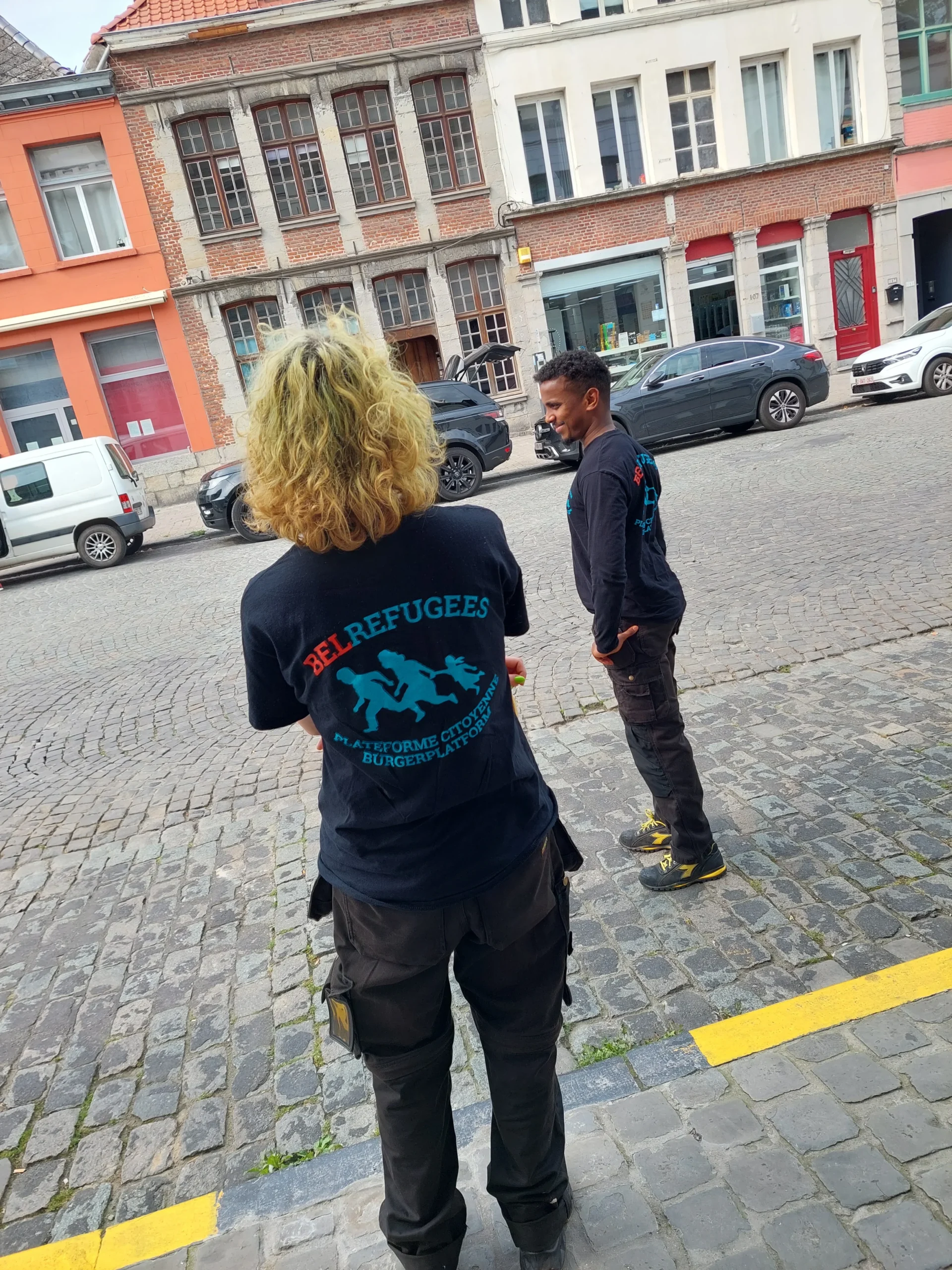đ€ BelRefugees 1 Femme blonde bouclĂ©e et homme cheveux brun court  de dos aborrant le logo