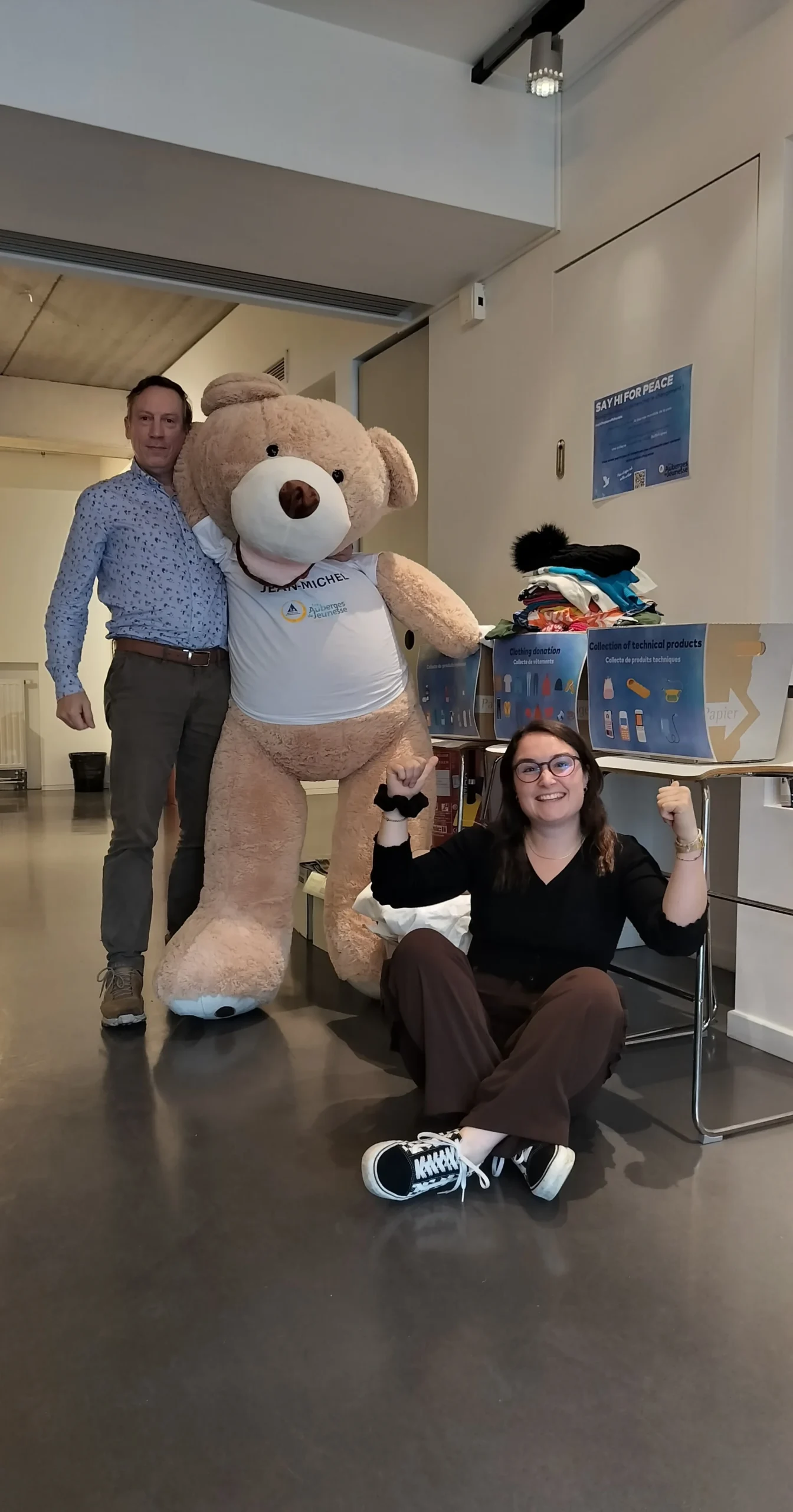 🕊️ Say Hi For Peace 2023 3 un homme debout tien tune énorme peluche ours dans les bras et une fille est assise parterre, souriante devant des boites de dons remplis